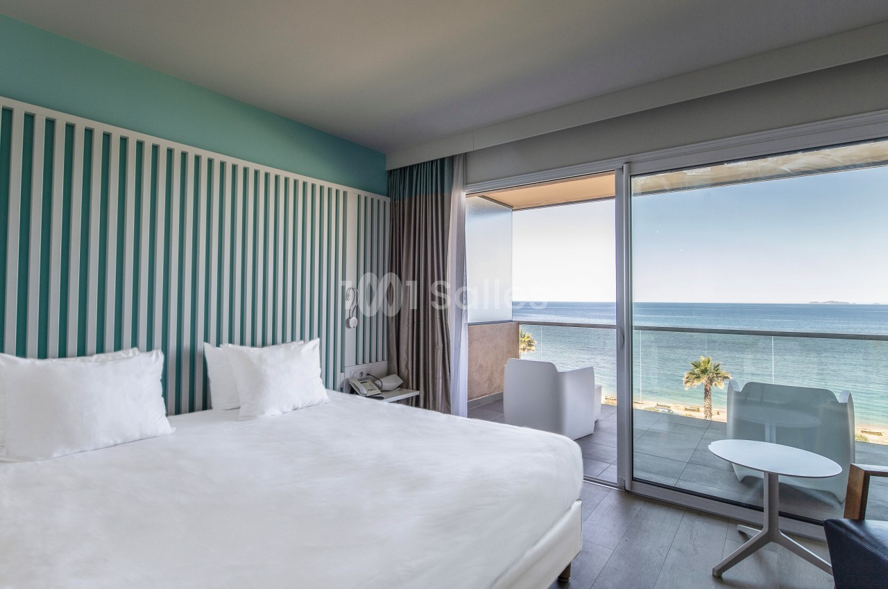 Chambre lumineuse avec lit double, décor moderne, balcon offrant une vue sur la mer et des palmiers.