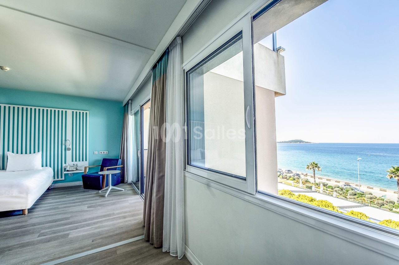 Chambre lumineuse avec vue sur la mer, grandes fenêtres, rideaux beiges et mobilier moderne.