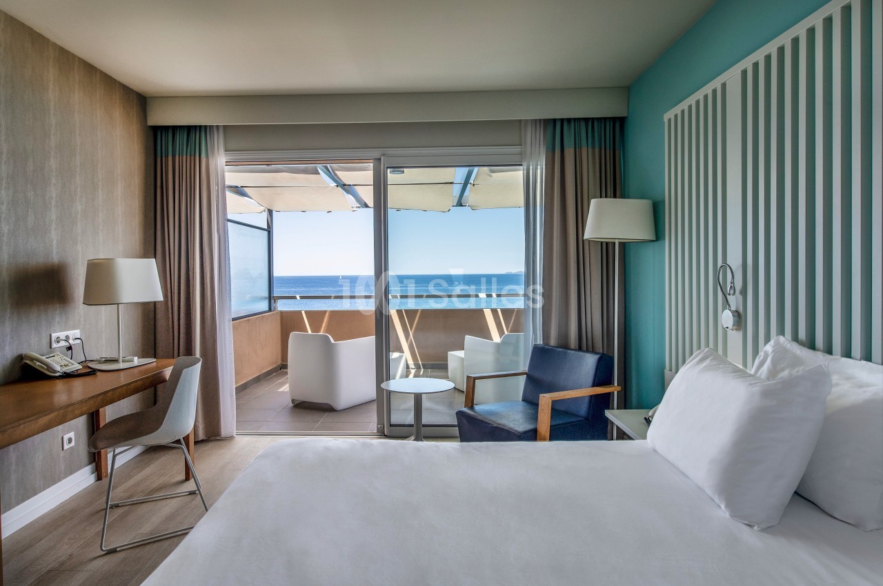 Chambre d'hôtel lumineuse avec lit, bureau, fauteuils et balcon offrant une vue sur la mer.