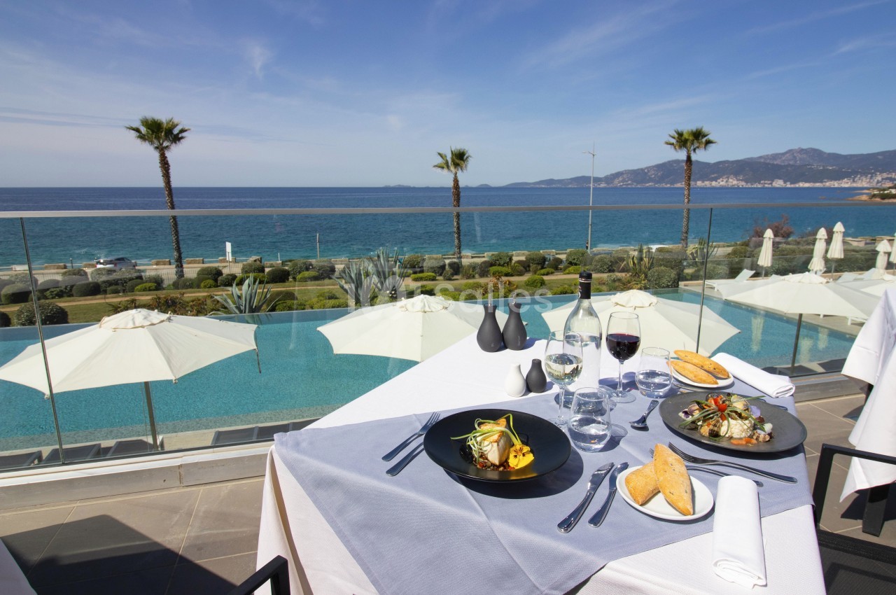 Table dressée avec des plats gastronomiques, vue sur une piscine, des parasols, des palmiers et la mer en arrière-plan.