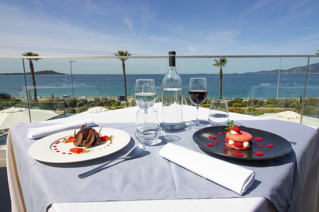 Table dressée avec vue sur la mer, comprenant des desserts élégants, un verre de vin rouge et une bouteille d'eau.