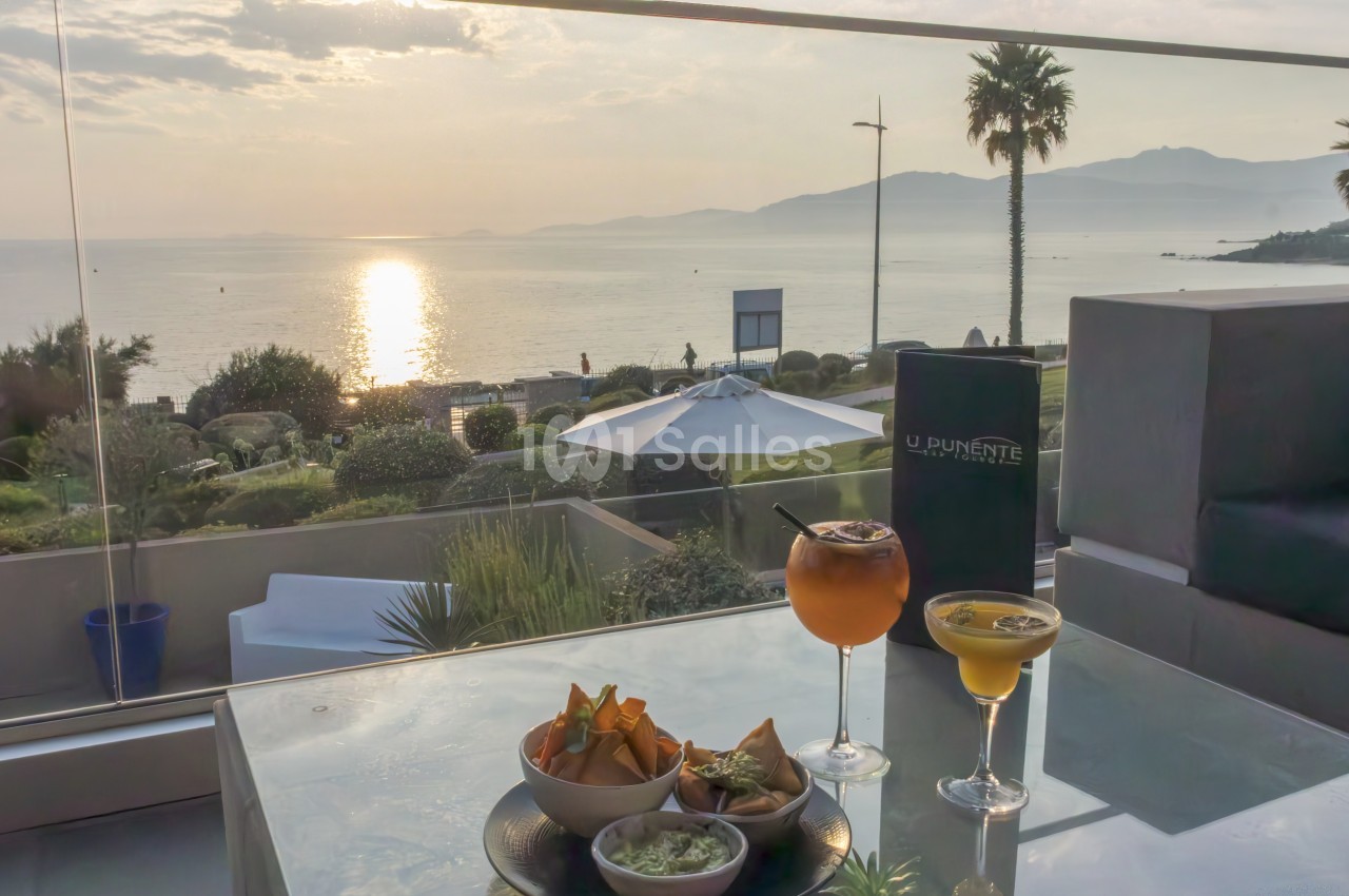 Deux cocktails et des amuse-bouches sur une table en terrasse avec vue sur la mer au coucher du soleil.