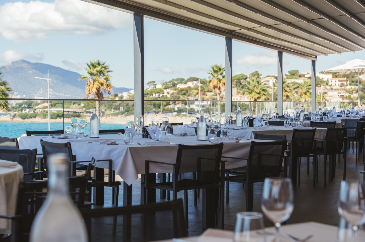 Terrasse d'un restaurant avec tables dressées, vue sur la mer, des palmiers et des collines en arrière-plan.