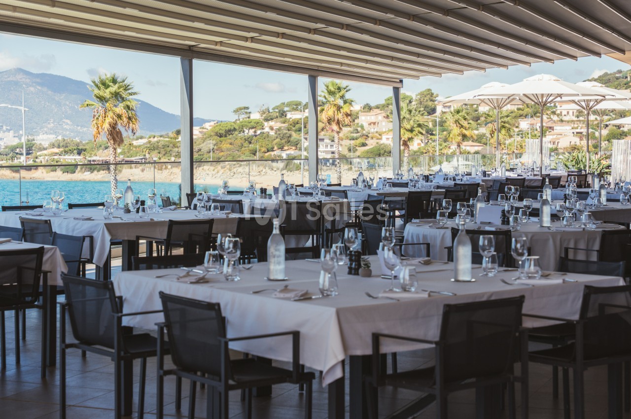 Salle de restaurant en terrasse avec vue sur la mer, tables dressées et paysage côtier en arrière-plan.
