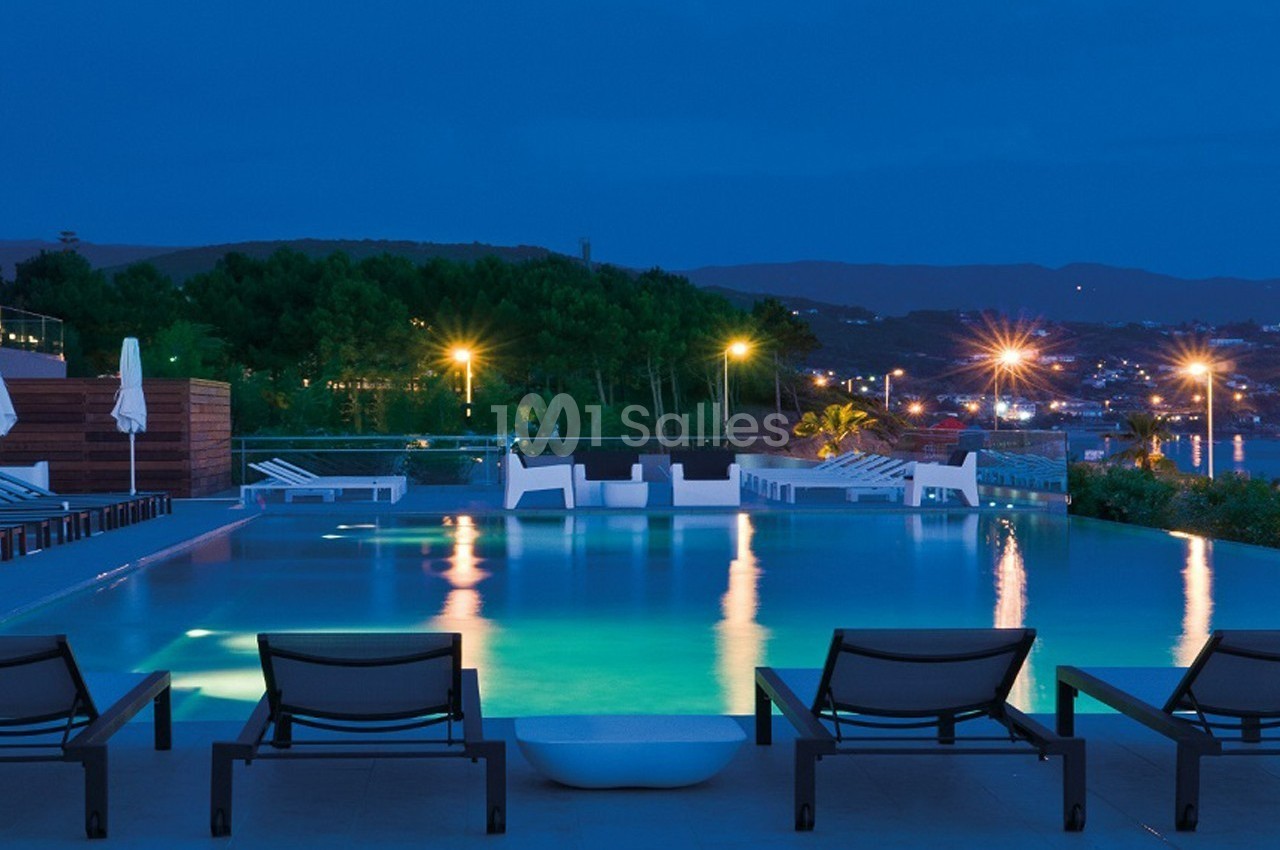 Piscine éclairée en soirée avec chaises longues au premier plan et vue sur une ville illuminée en arrière-plan.