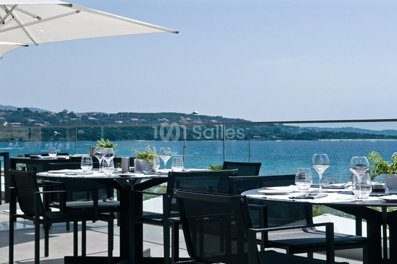 Terrasse d'un restaurant avec tables dressées, vue sur la mer et collines en arrière-plan par temps ensoleillé.