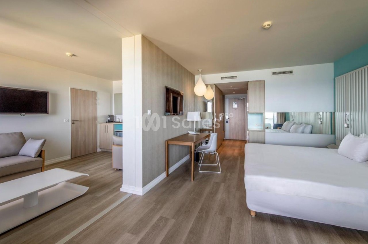 Chambre d'hôtel moderne avec lit double, coin salon, bureau, télévision murale et sol en parquet clair.