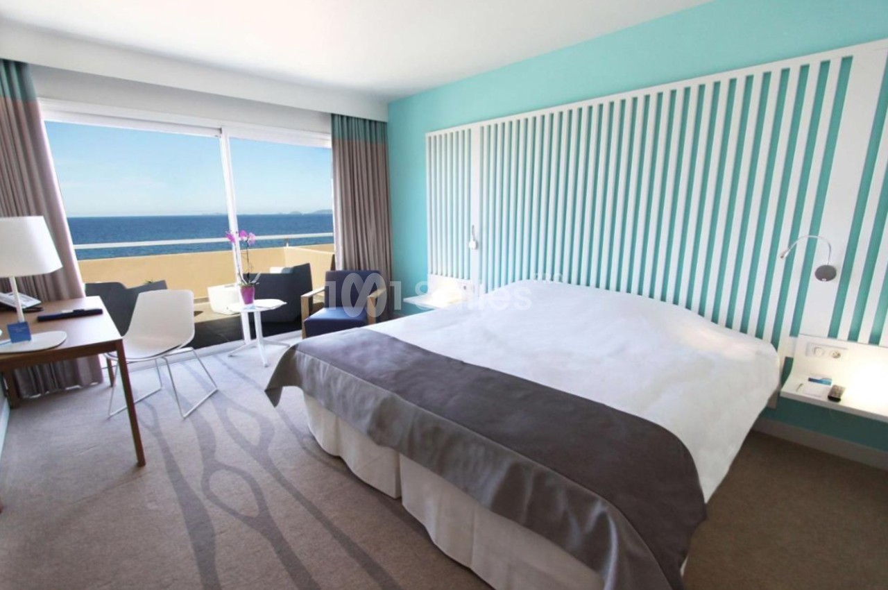 Chambre lumineuse avec lit double, bureau, fauteuils, baie vitrée donnant sur une terrasse et vue sur la mer.