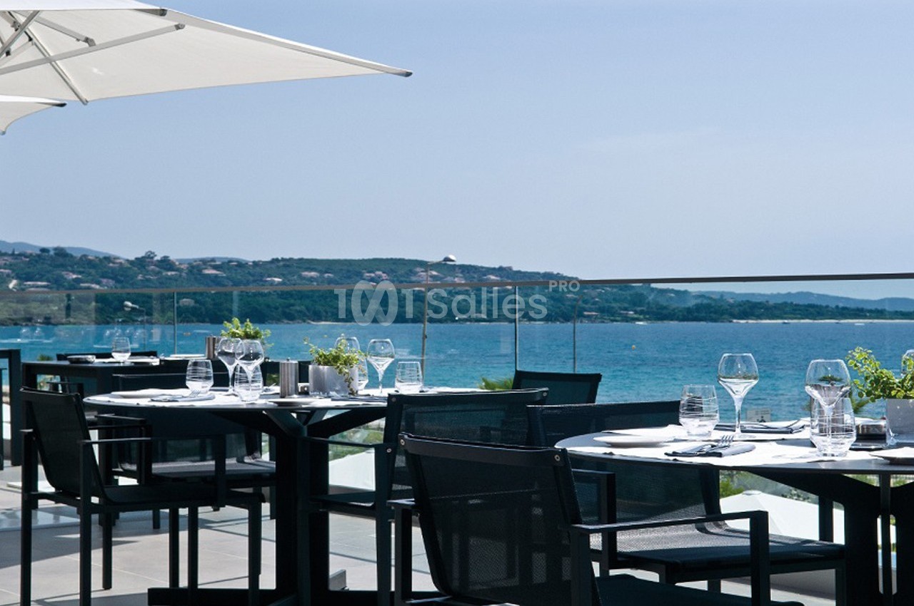 Terrasse d'un restaurant avec tables dressées, vue sur la mer et collines en arrière-plan par temps ensoleillé.