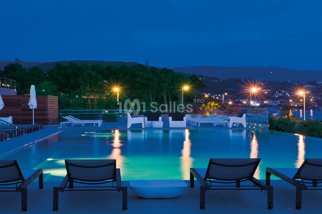 Piscine extérieure éclairée de nuit avec transats au premier plan et vue sur une ville illuminée en arrière-plan.