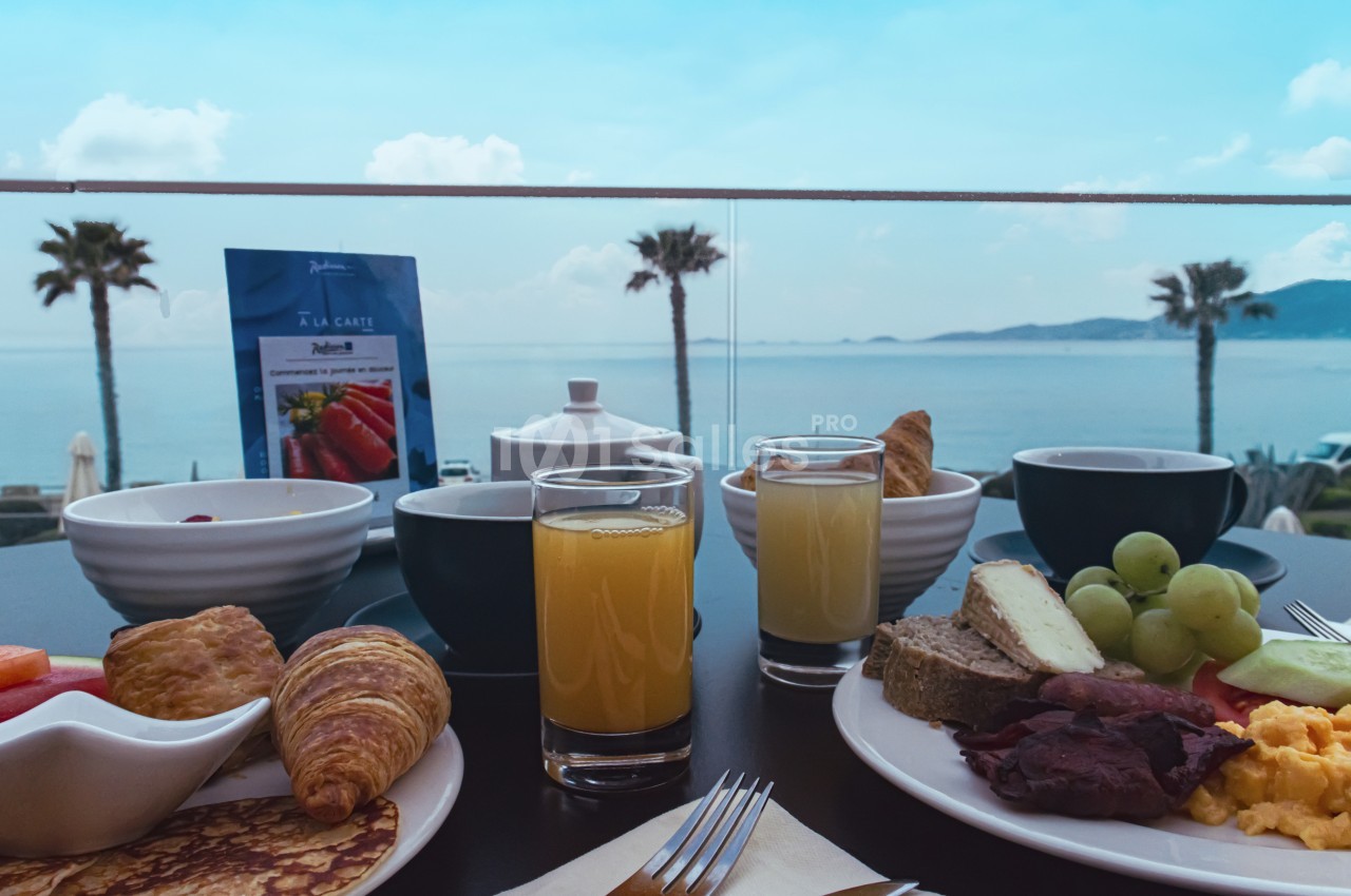 Petit-déjeuner varié avec fruits, viennoiseries et jus d'orange, servi en terrasse avec vue sur la mer et des palmiers.