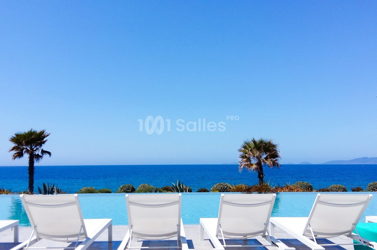 Chaises longues blanches alignées face à une piscine à débordement avec vue sur la mer et deux palmiers.
