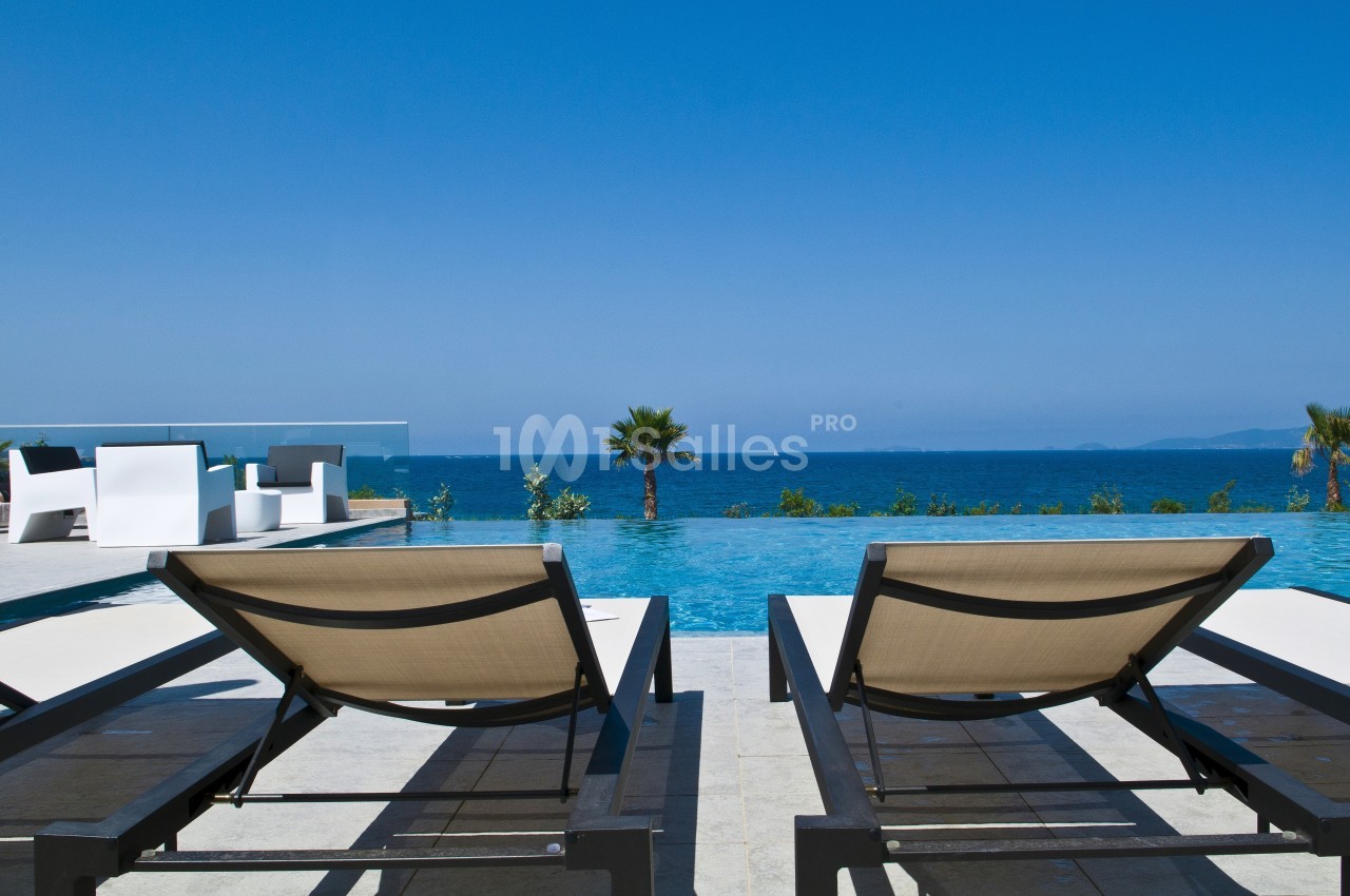 Deux transats face à une piscine à débordement avec vue sur la mer et un ciel bleu dégagé.