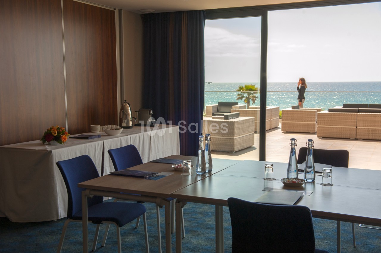 Salle de réunion lumineuse avec vue sur une terrasse donnant sur la mer, équipée de tables, chaises et boissons.