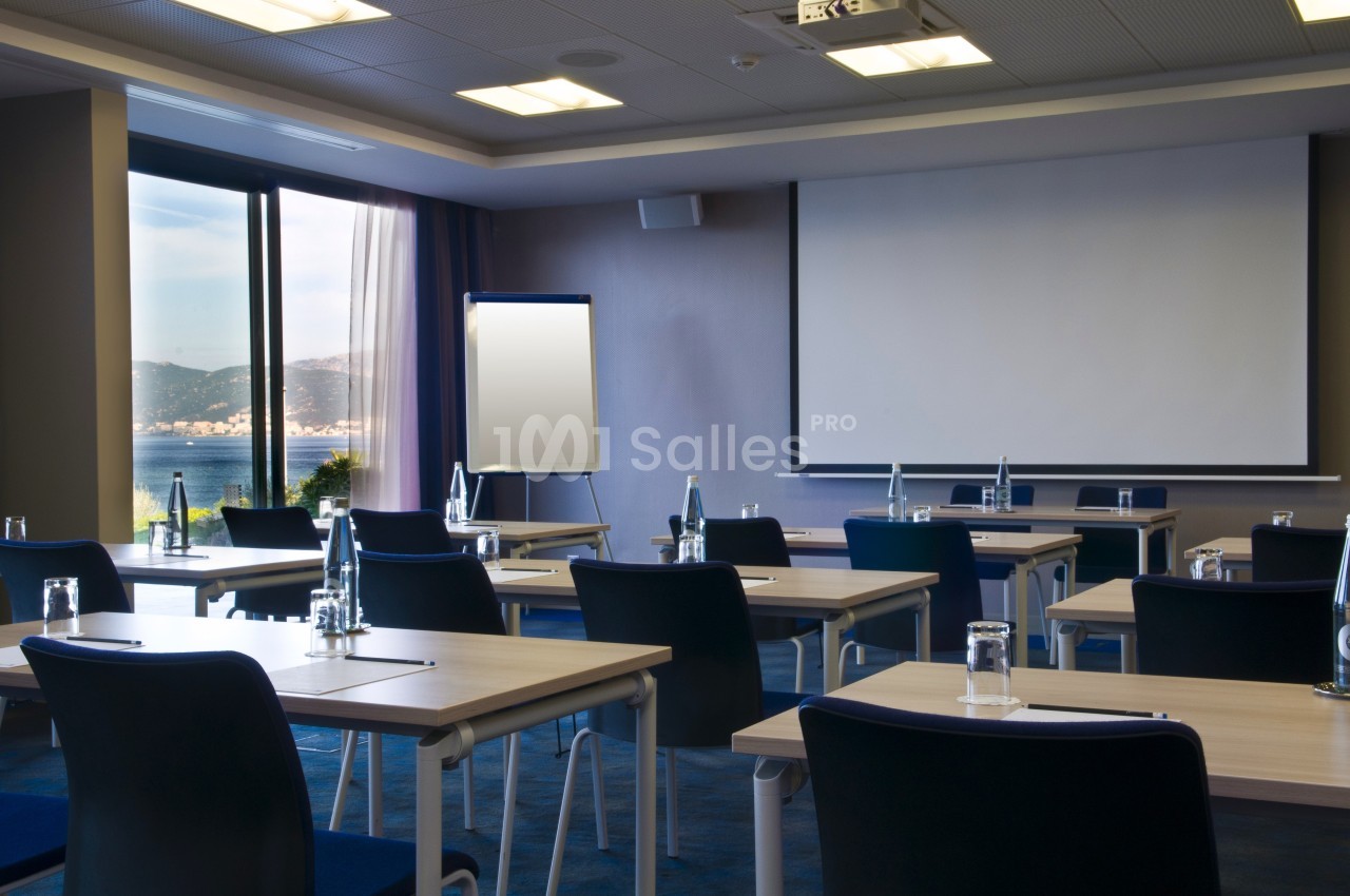 Salle de réunion lumineuse avec tables, chaises, écran de projection, tableau blanc et vue sur la mer.