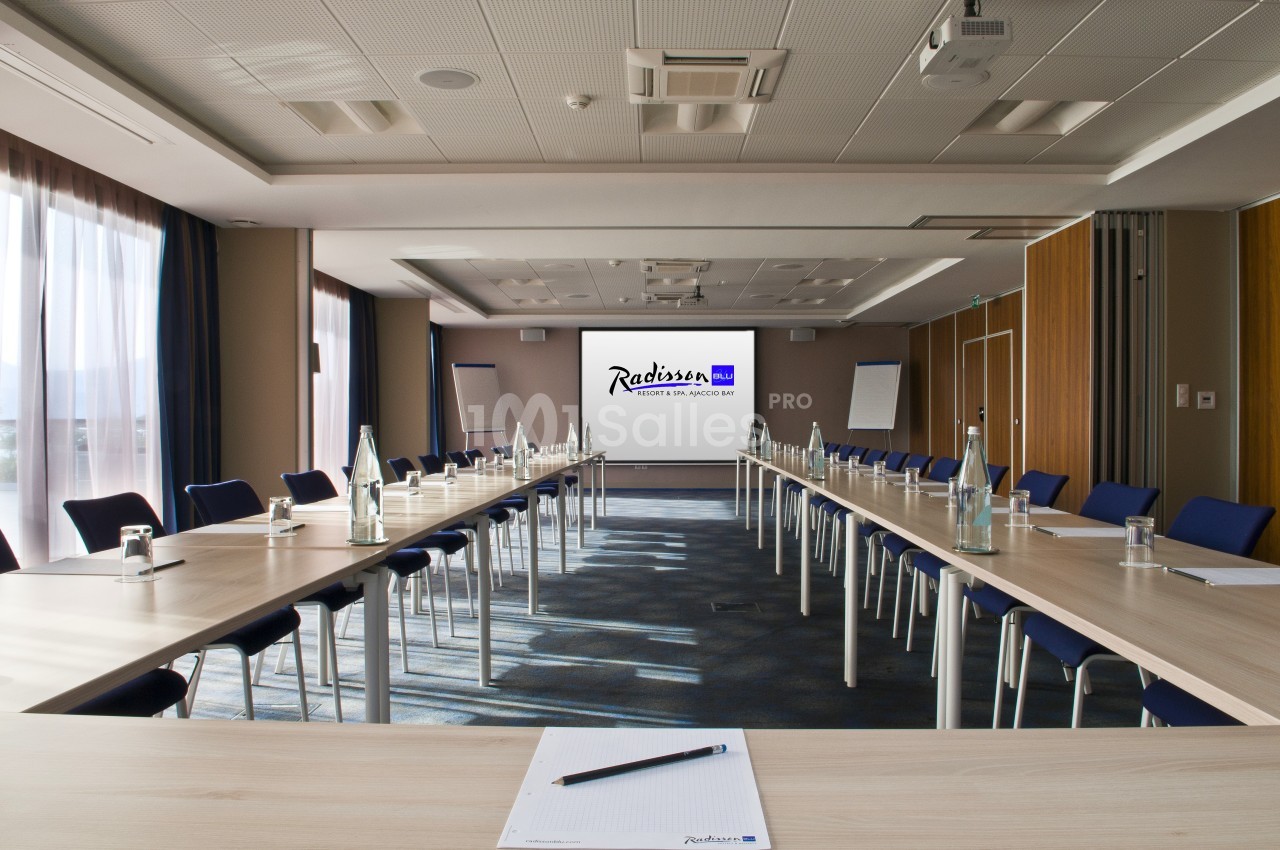 Salle de réunion lumineuse avec tables disposées en U, chaises bleues et écran de présentation au fond.