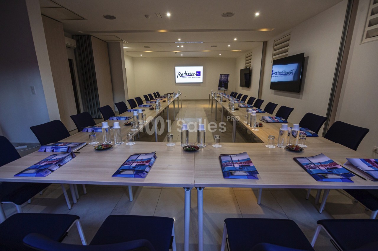 Salle de réunion moderne avec tables disposées en U, chaises, documents, bouteilles d'eau et écran allumé en fond.