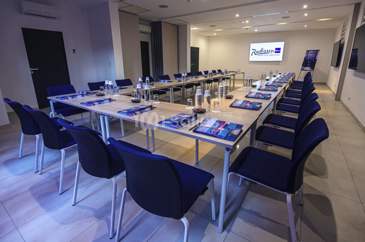 Salle de réunion moderne avec tables disposées en U, chaises bleues et matériel de conférence sur les tables.