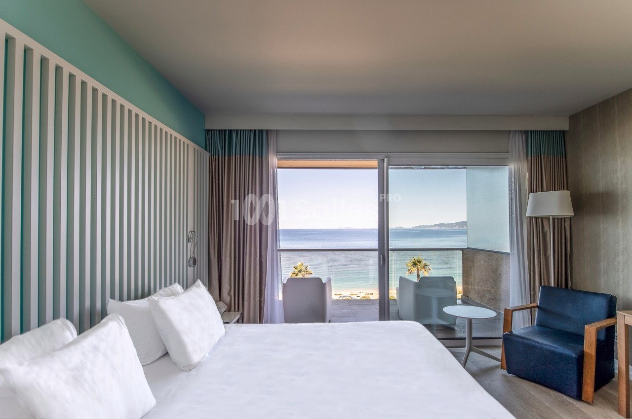 Chambre d'hôtel lumineuse avec lit double, fauteuil, balcon et vue sur la mer.