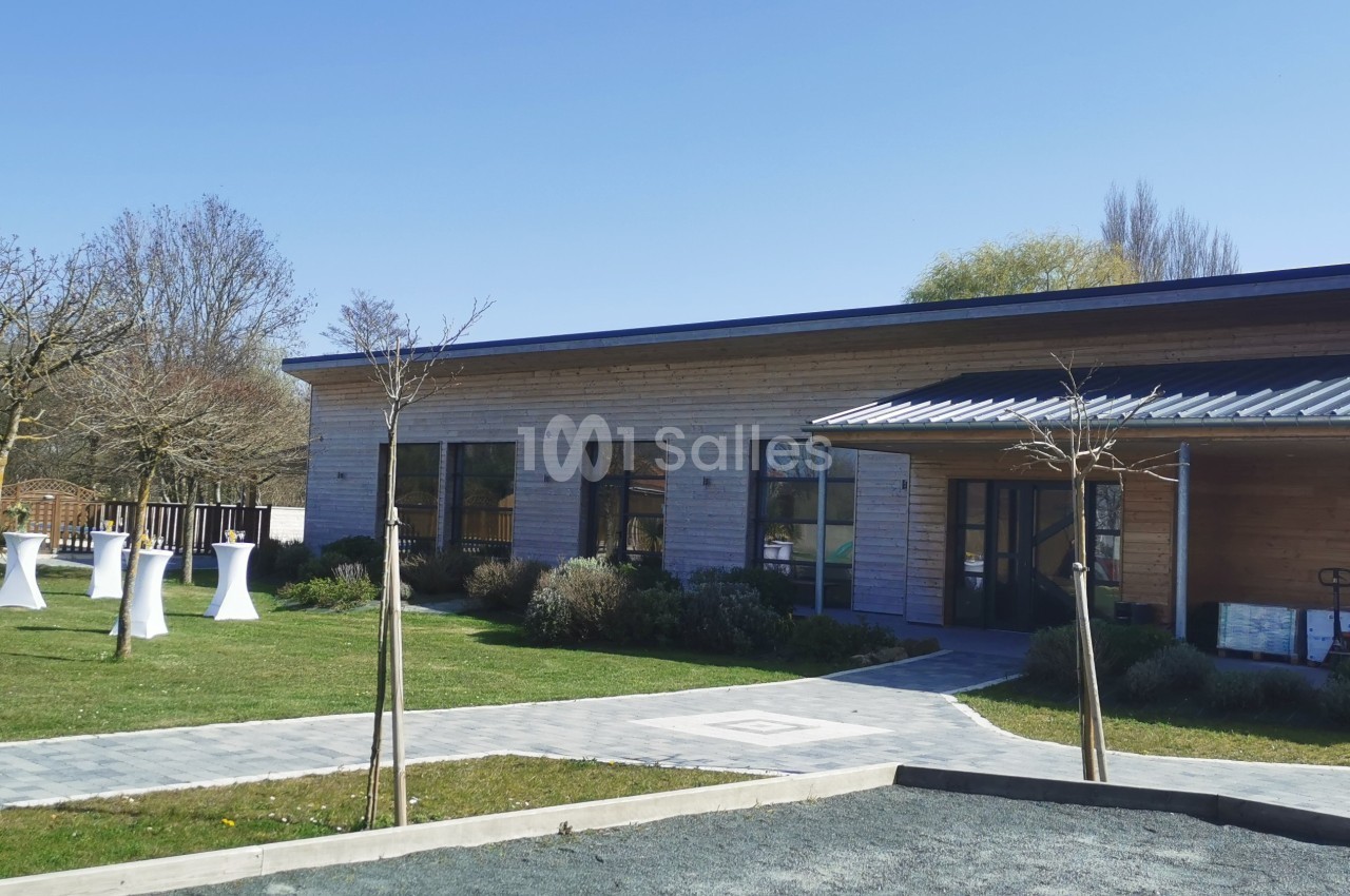 Bâtiment moderne en bois entouré d'un jardin avec des arbres, des tables hautes blanches et un chemin pavé.