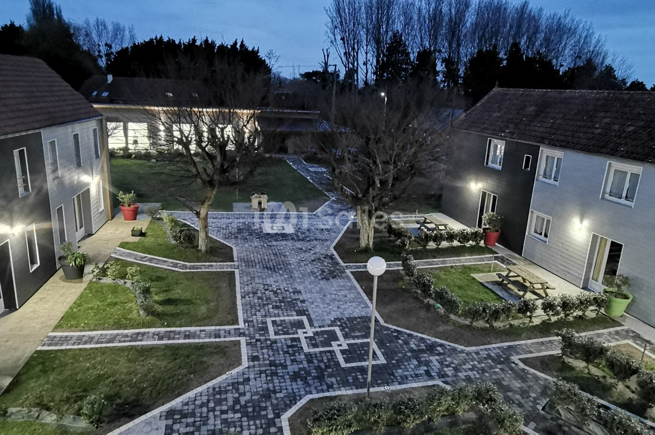 Cour pavée entourée de bâtiments modernes avec espaces verts, arbres et éclairage extérieur au crépuscule.