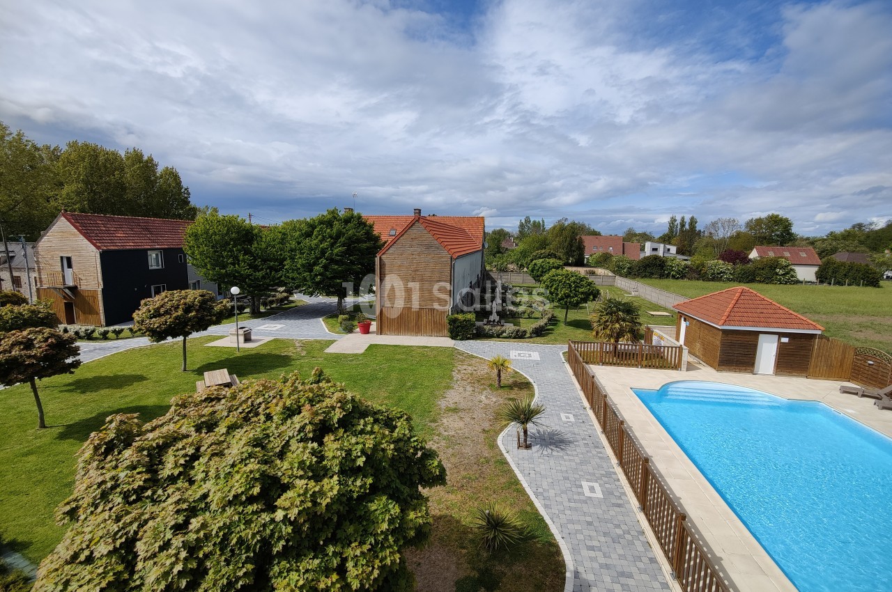 Vue d'un jardin avec pelouse, arbres, piscine entourée de dalles et maisons aux toits rouges sous un ciel partiellement…