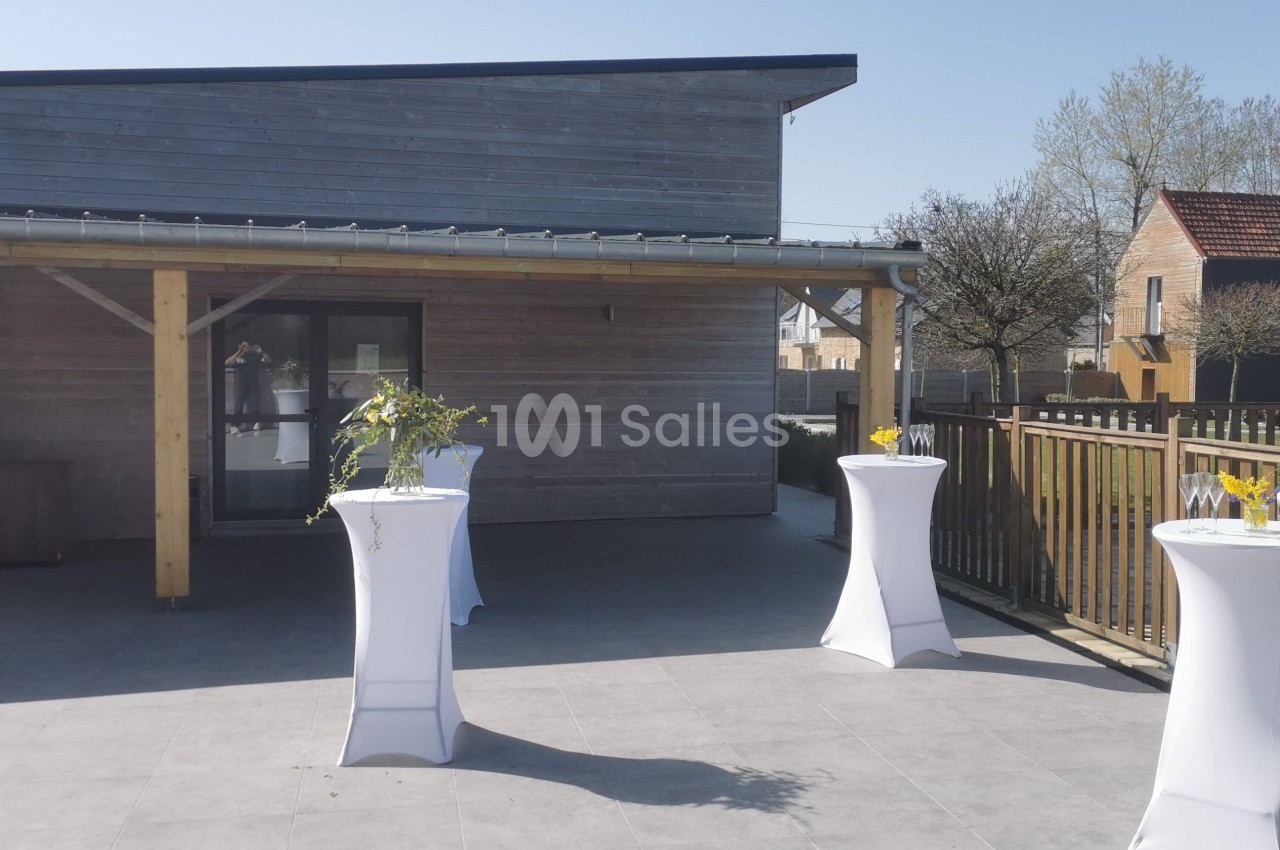 Terrasse ensoleillée avec tables hautes drapées de blanc, décorées de fleurs et de vases, devant un bâtiment en bois.