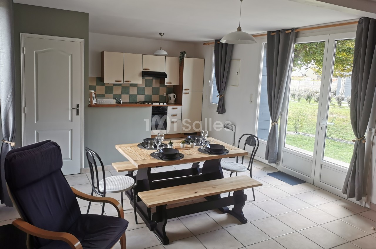 Cuisine ouverte avec table en bois dressée, chaises, fauteuil, et vue sur un jardin à travers des portes vitrées.