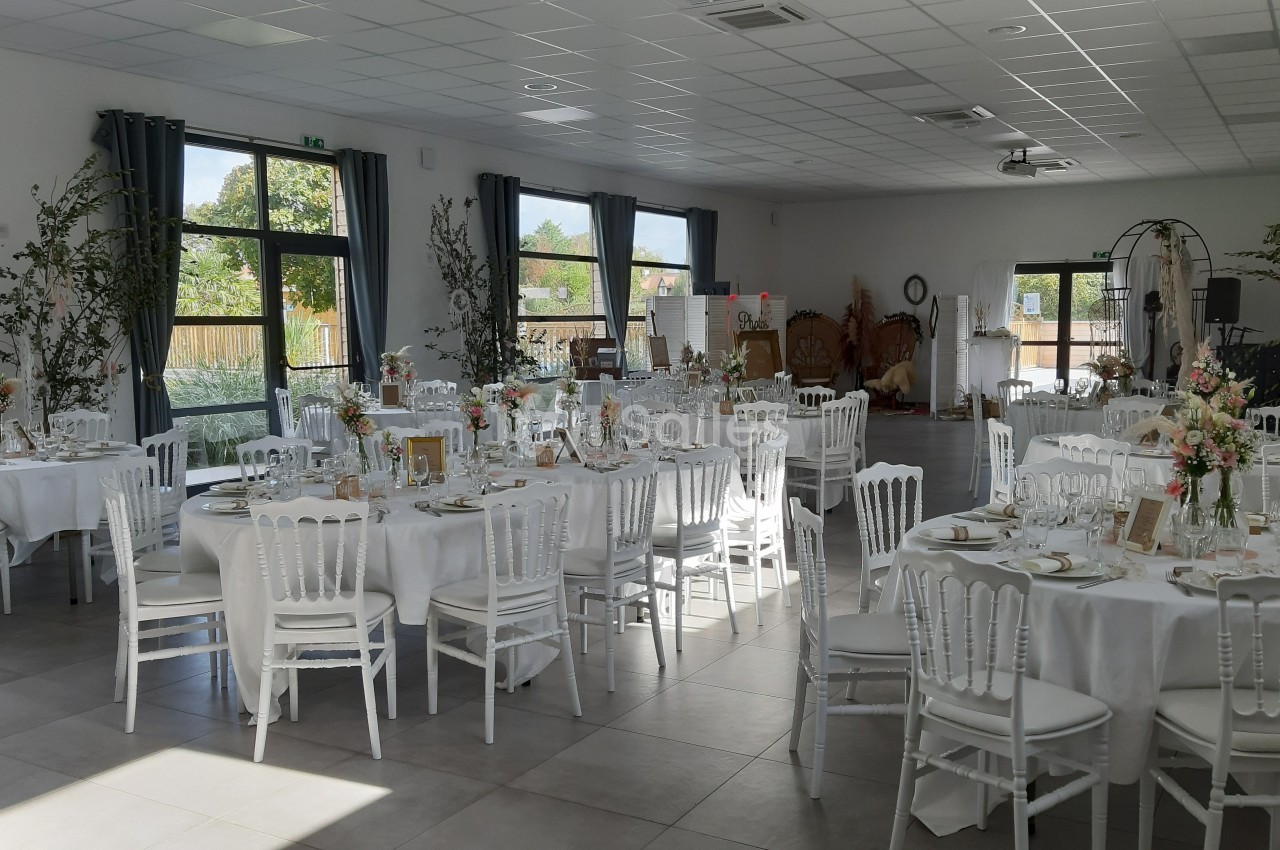 Salle de réception lumineuse avec tables décorées de nappes blanches, chaises élégantes et arrangements floraux.
