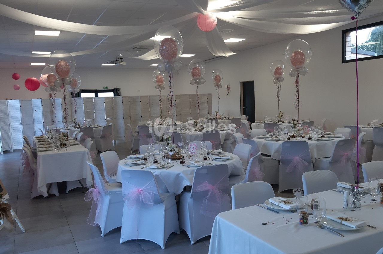 Salle de réception décorée avec des tables rondes et rectangulaires, nappes blanches, ballons et rubans roses.