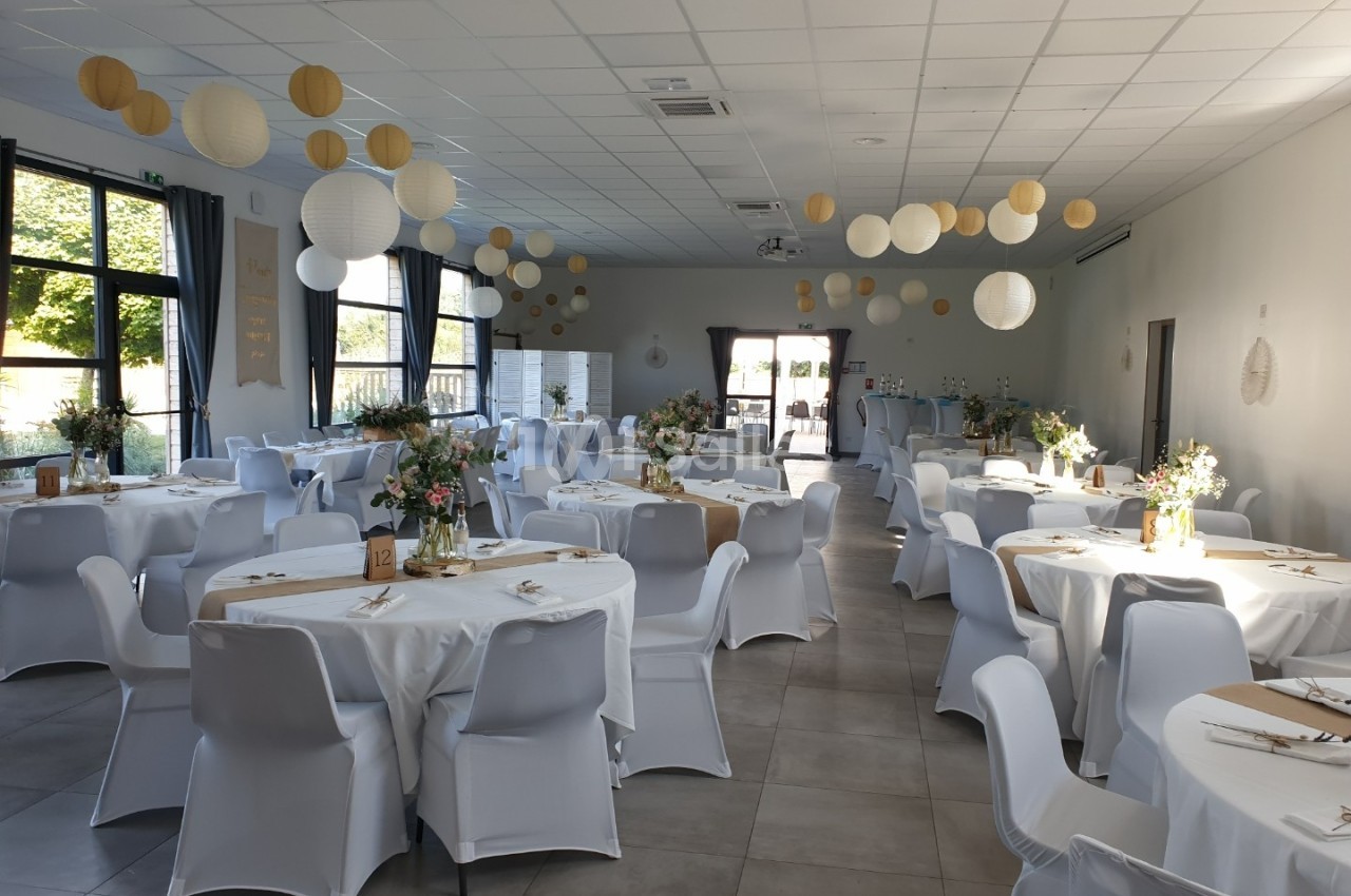Salle de réception décorée avec des tables rondes, nappes blanches, fleurs et lanternes suspendues.