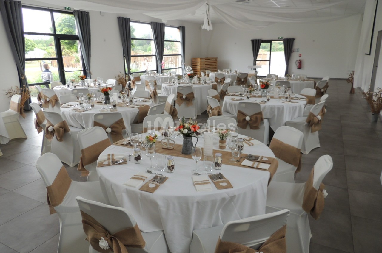 Salle de réception décorée avec des tables rondes, nappes blanches, chaises ornées de nœuds en toile de jute.