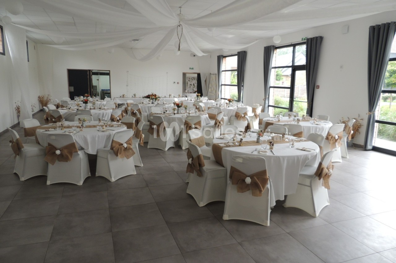 Salle de réception décorée avec des tables rondes, nappes blanches et chaises ornées de nœuds en tissu beige.