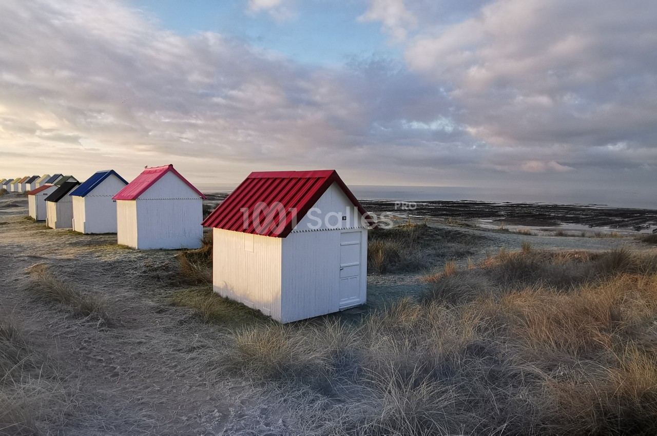 Alignement de petites cabanes blanches aux toits colorés sur une dune herbeuse, avec vue sur la mer et le ciel nuageux.