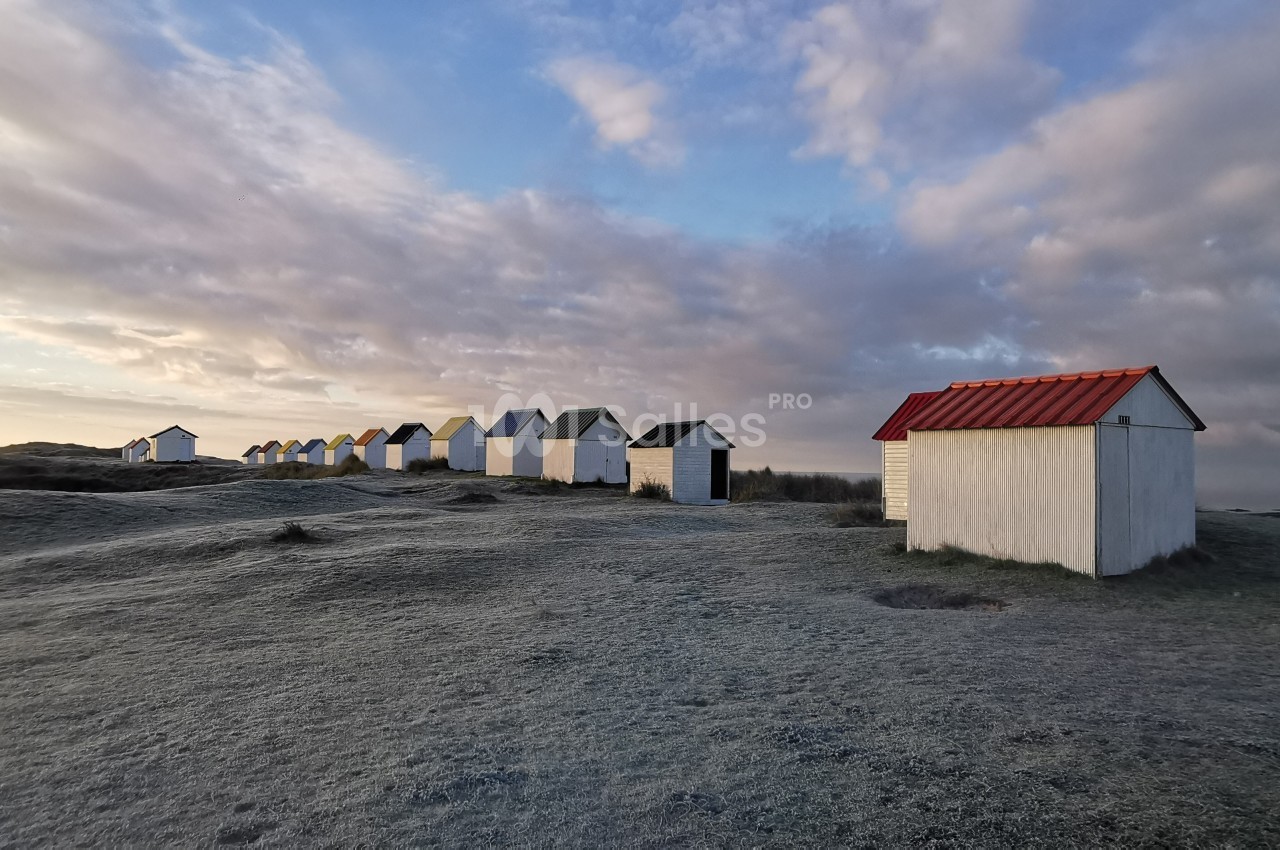 Alignement de petites cabanes blanches sur un terrain herbeux givré, sous un ciel nuageux au lever du jour.