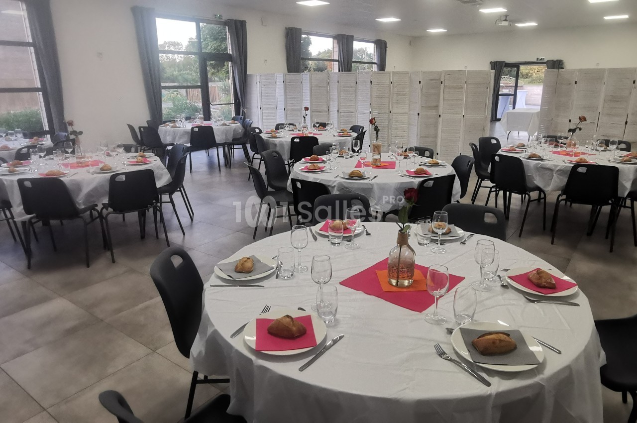 Salle de réception avec tables rondes dressées, nappes blanches, serviettes roses et vaisselle, prête pour un événement.