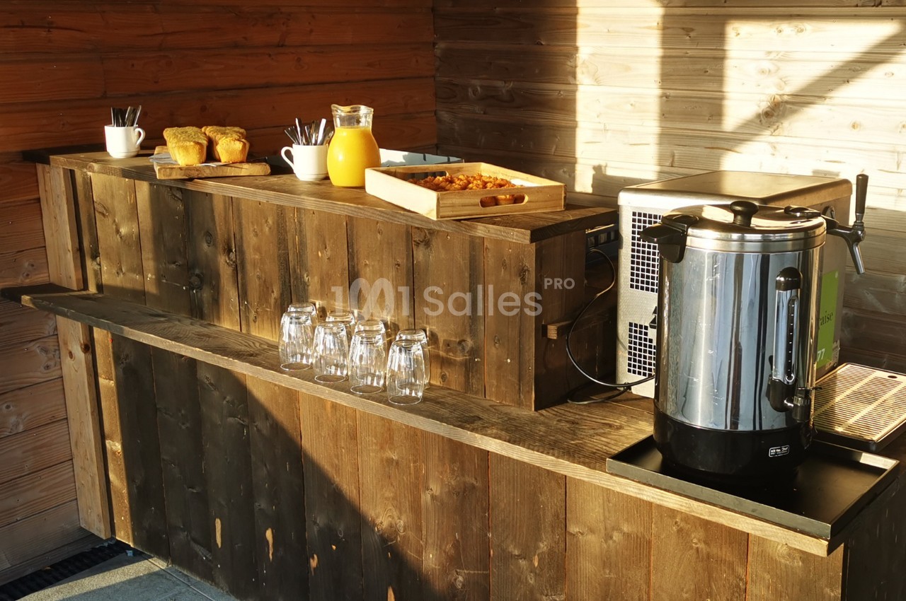 Buffet en bois avec cafetière, jus d'orange, viennoiseries, tasses et verres, éclairé par la lumière naturelle.