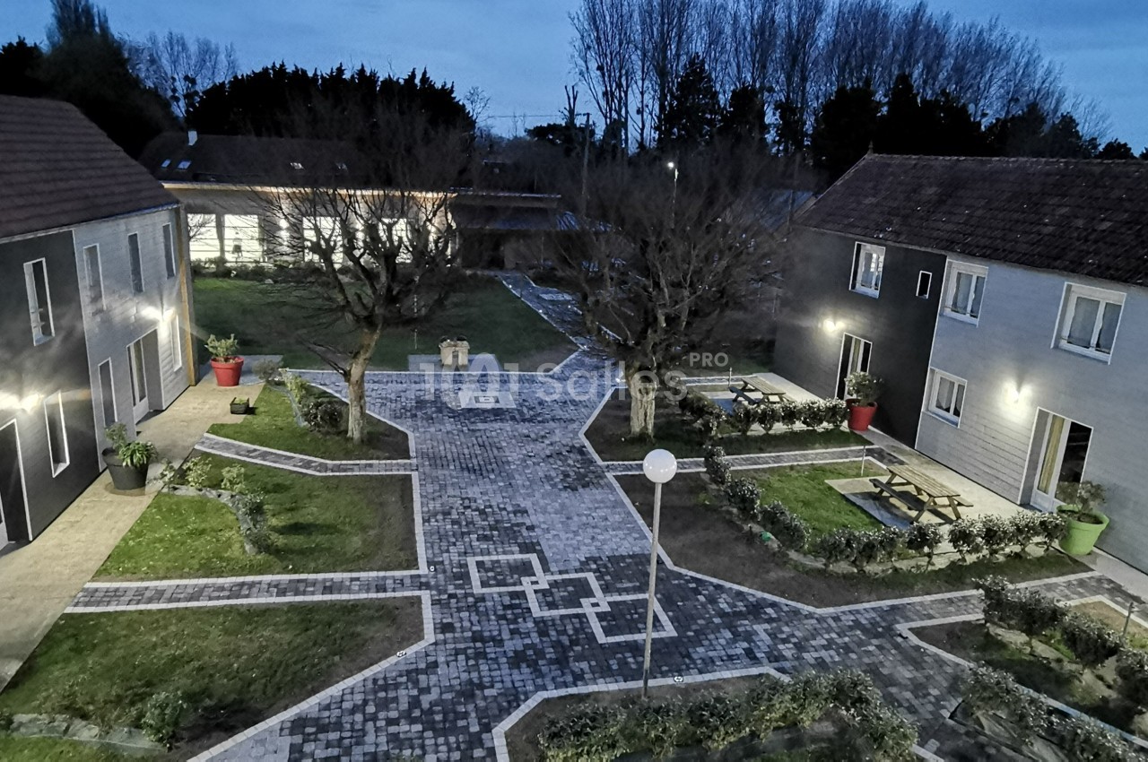 Cour pavée entourée de bâtiments modernes avec espaces verts, arbres et éclairage extérieur au crépuscule.