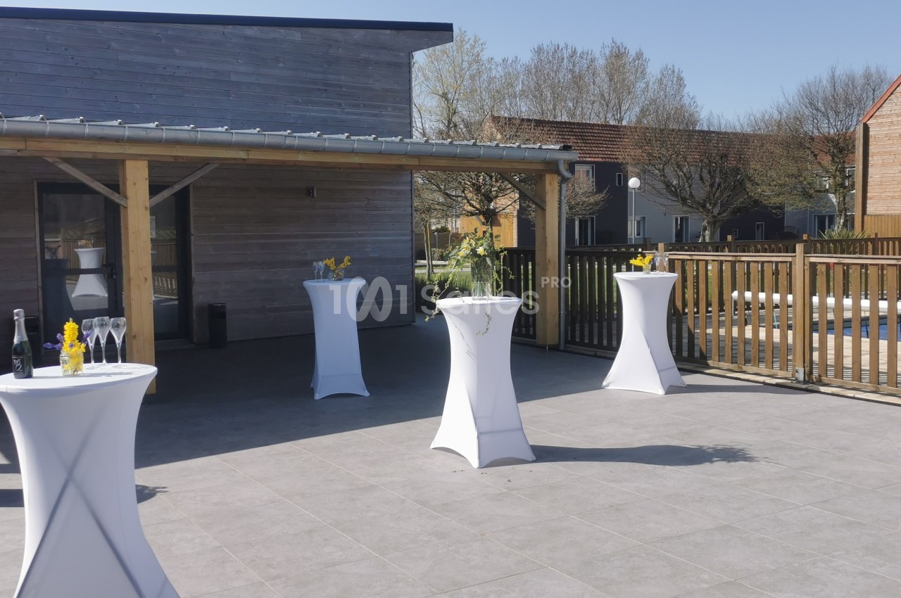 Tables hautes drapées de blanc disposées en extérieur sur une terrasse ensoleillée, avec quelques décorations florales.