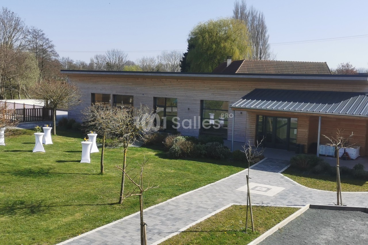 Bâtiment moderne en bois entouré d'un jardin avec des arbres et des tables hautes blanches en extérieur.