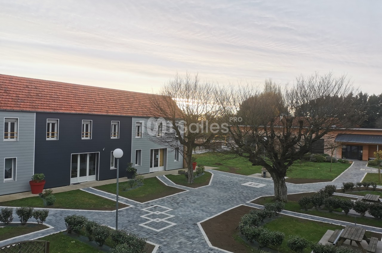 Vue d'une cour pavée avec bâtiments modernes, arbres taillés et bancs, sous un ciel partiellement nuageux.