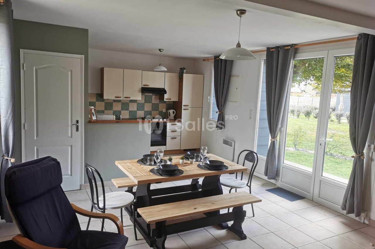 Salle à manger avec table en bois, chaises, cuisine ouverte et grandes fenêtres donnant sur un jardin.