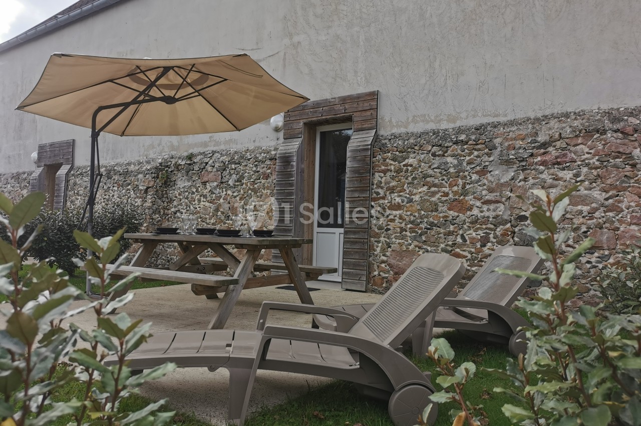 Table de pique-nique et chaises longues disposées sur une terrasse en béton devant un mur en pierre, avec un parasol beige.