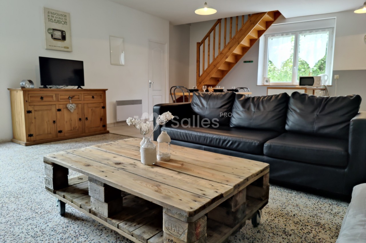 Salon lumineux avec canapé en cuir noir, table basse en palettes, meuble en bois, escalier et fenêtre donnant sur un jardin.