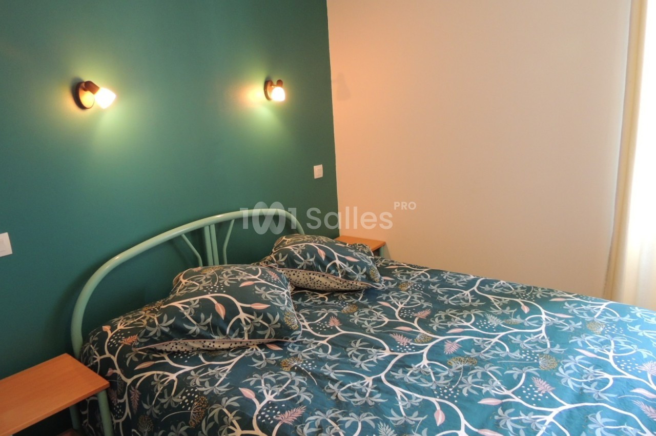 Lit double avec une tête de lit en métal vert, draps à motifs floraux, éclairé par deux appliques murales.