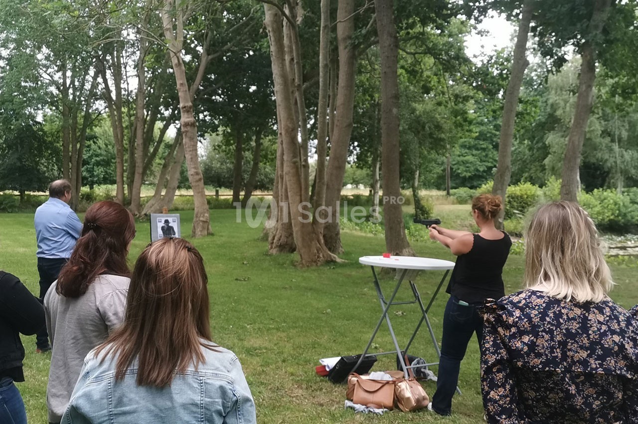 Une femme tire avec une arme à air comprimé sur une cible dans un parc, observée par plusieurs personnes.
