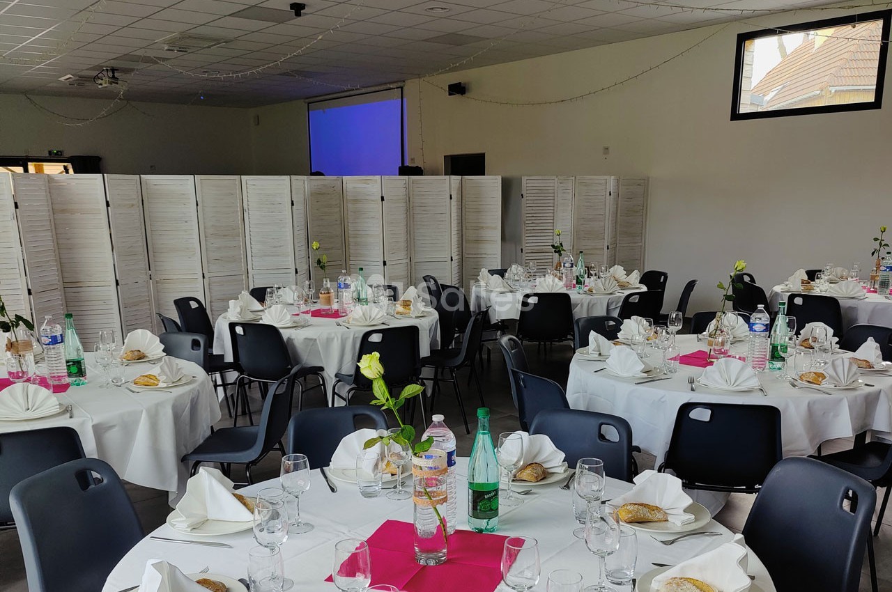 Salle de réception avec tables rondes dressées, nappes blanches, serviettes pliées, fleurs et bouteilles d'eau.