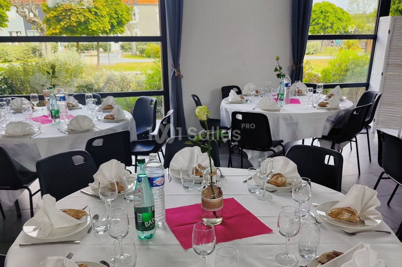 Salle de réception avec tables rondes dressées, nappes blanches, serviettes pliées, verres, et décoration florale.