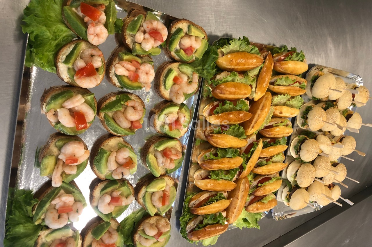 Plateaux de canapés variés avec crevettes, avocat, petits pains garnis et mini-burgers sur une table en inox.