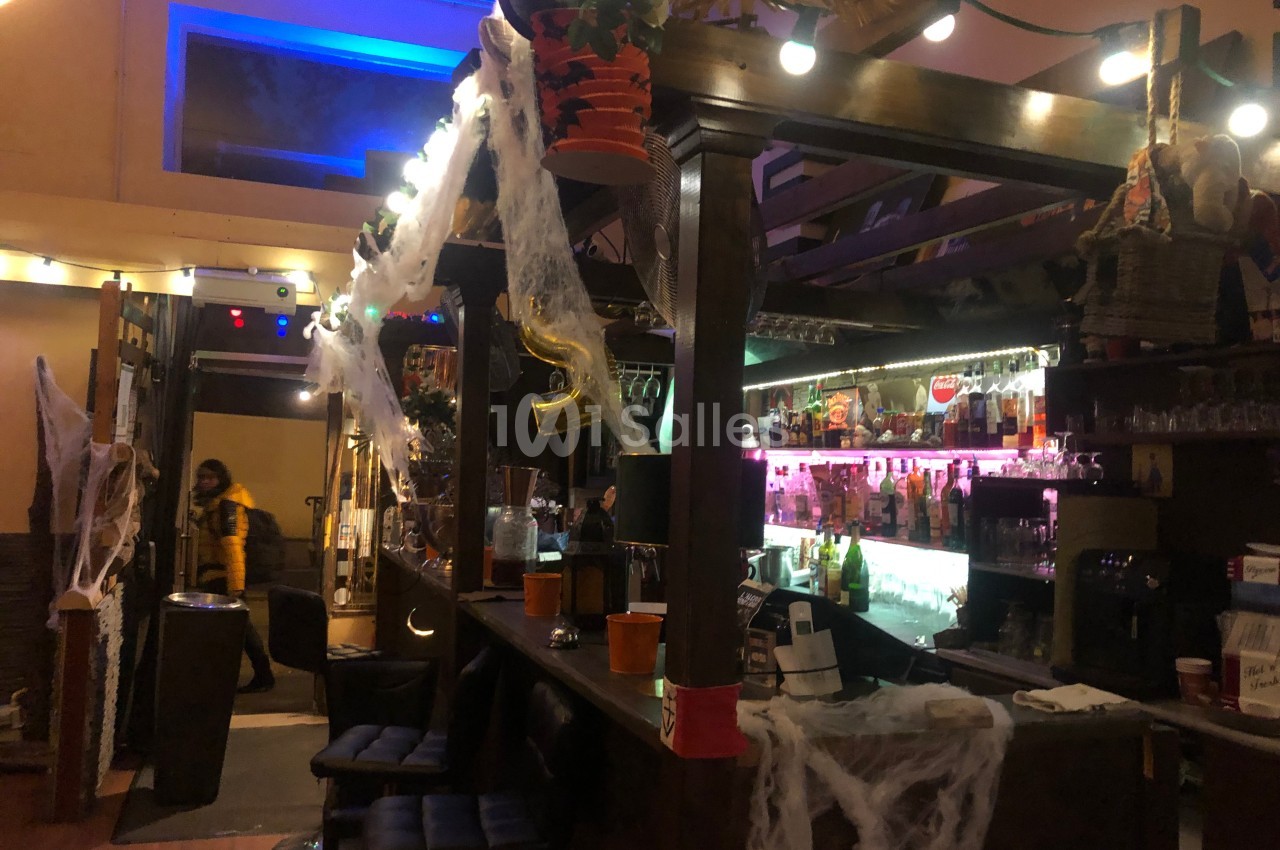 Bar décoré pour Halloween avec des toiles d'araignée, des lumières colorées et des bouteilles alignées sur les étagères.